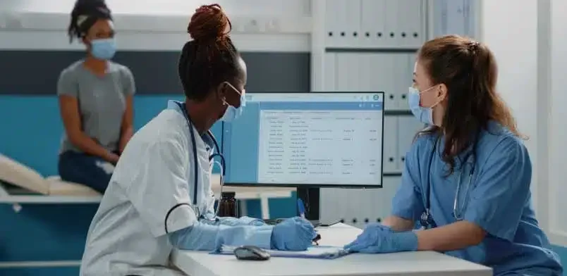 6-Effective-Ways-To-AR-Days-in-Oncology-Billing-imge Two women in medical scrubs are seated at a desk, discussing and working on paperwork together.